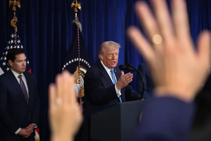 Donald Trump vorbește la o conferință, cu mâini ridicate în fundal.