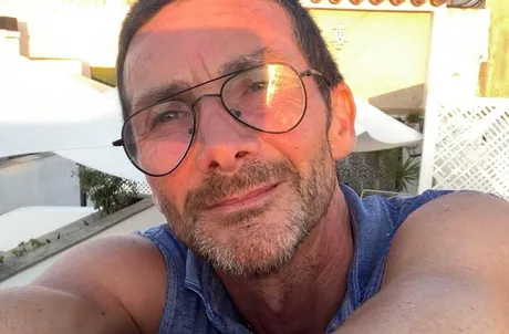 Un bărbat zâmbește în fața camerei, purtând o bluză fără mâneci.