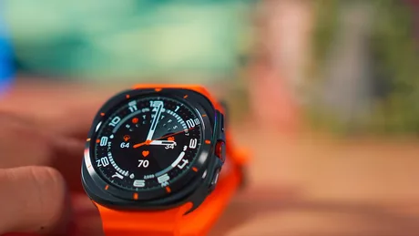 Ceas inteligent cu display colorat și curea portocalie, pe un fundal neclar.