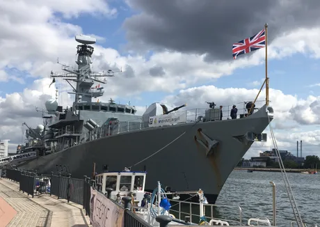 Nava militară britanică ancorată în port, cu steagul Regatului Unit fluturând.