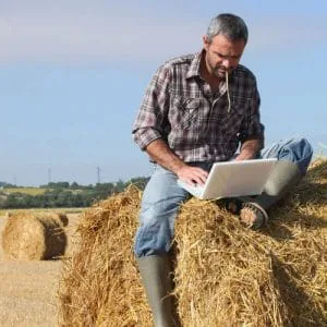 Fermier lucrând pe laptop în câmp, pe un balot de fân.