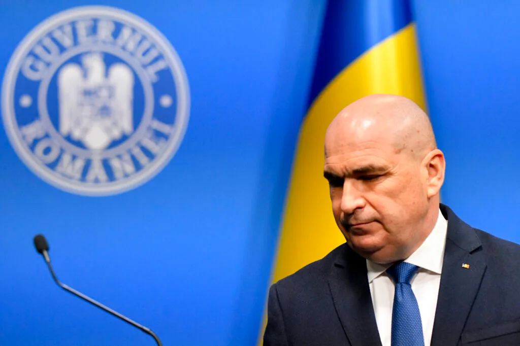 Ilie Bolojan în fața unui banner guvernamental, reflectând tensiunea politică actuală.
