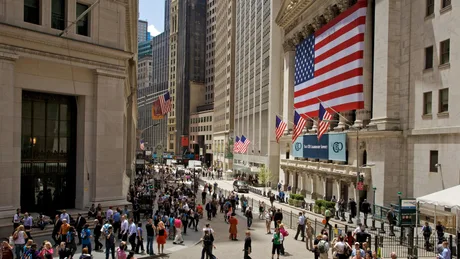 Străzi aglomerate pe Wall Street, cu clădiri înalte și steaguri americane.