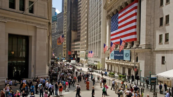 Străzi aglomerate pe Wall Street, cu clădiri înalte și steaguri americane.