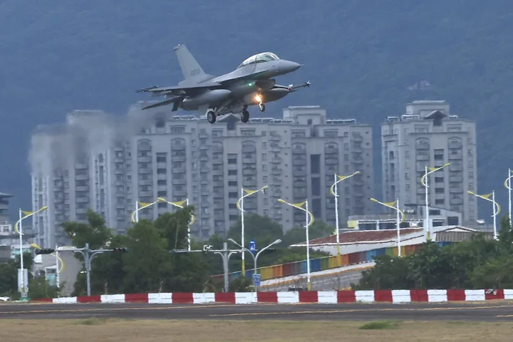 Avion F-16V în zbor, cu clădiri moderne în fundal.