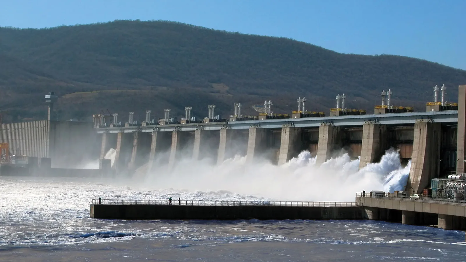Barajul Hidroelectrica cu ape învolburate și peisaj montan în fundal.