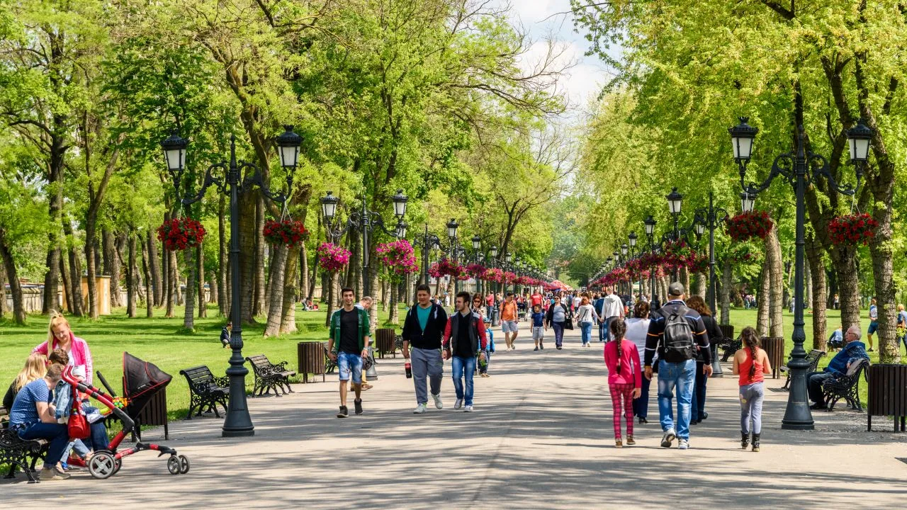 Oameni plimbându-se printr-un parc verde, cu copaci și flori colorate.