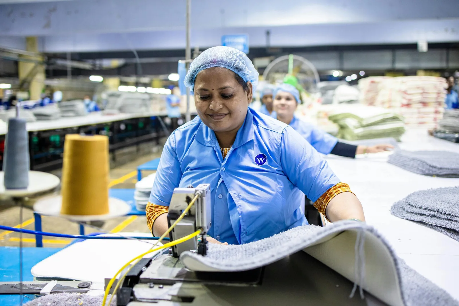 Femeie lucrând la o mașină de cusut într-o fabrică, zâmbind.