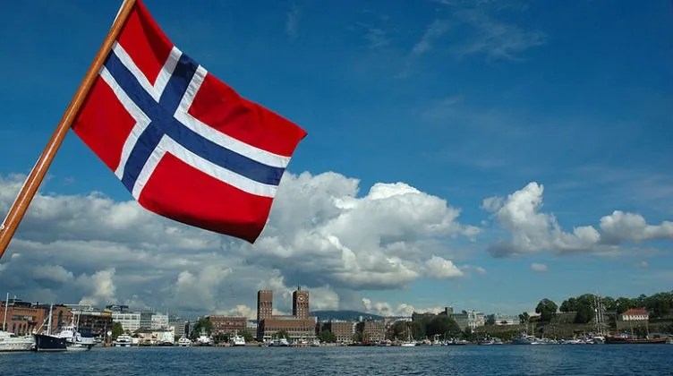 Steagul Norvegiei fluturând pe malul unui lac, cu cer senin.