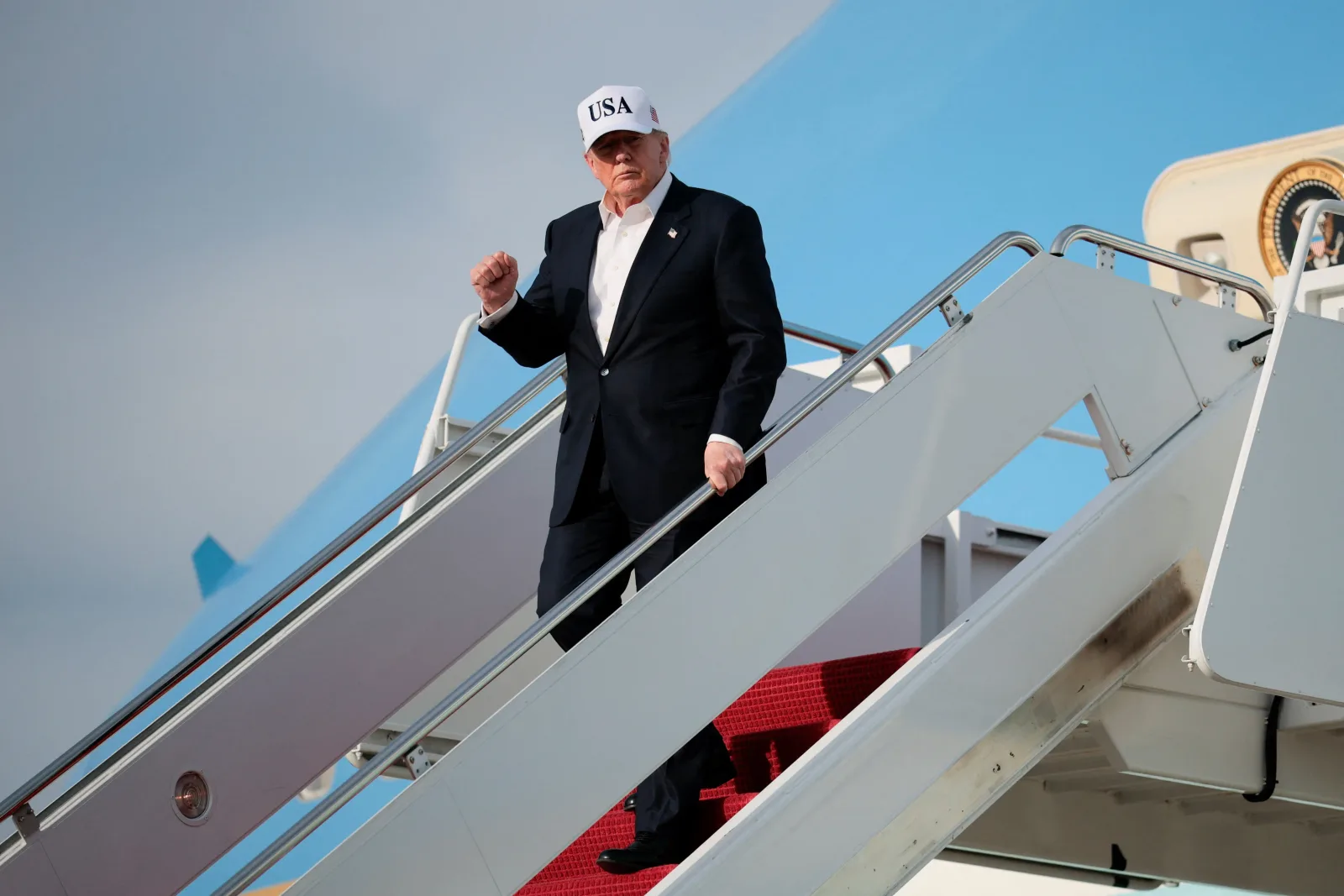 Donald Trump coboară dintr-un avion, purtând o șapcă albă cu inscripția SUA.
