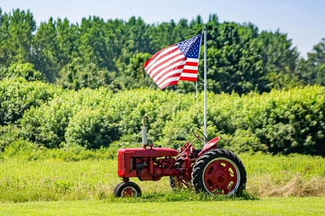 Tractor roșu pe câmp cu steagul american în fundal.
