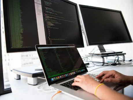 Un programator lucrează la un laptop, cu două monitoare în fundal.