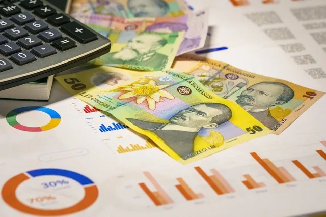 Bancnote românești și un calculator pe un birou cu grafice financiare.