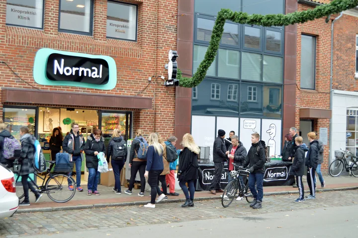 Oameni la intrarea unui magazin Normal, atmosfera animată pe stradă.