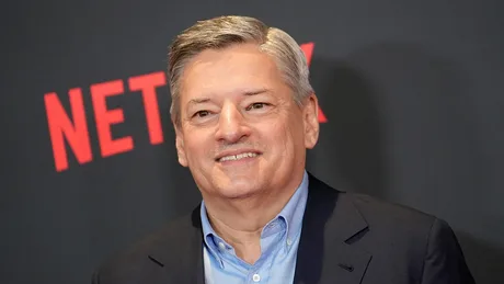 Ted Sarandos zâmbind la un eveniment Netflix, cu logo-ul companiei în fundal.