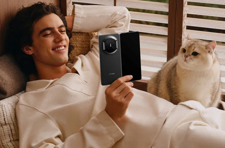 Un tânăr relaxat, folosind un telefon pliabil, alături de o pisică pe canapea.