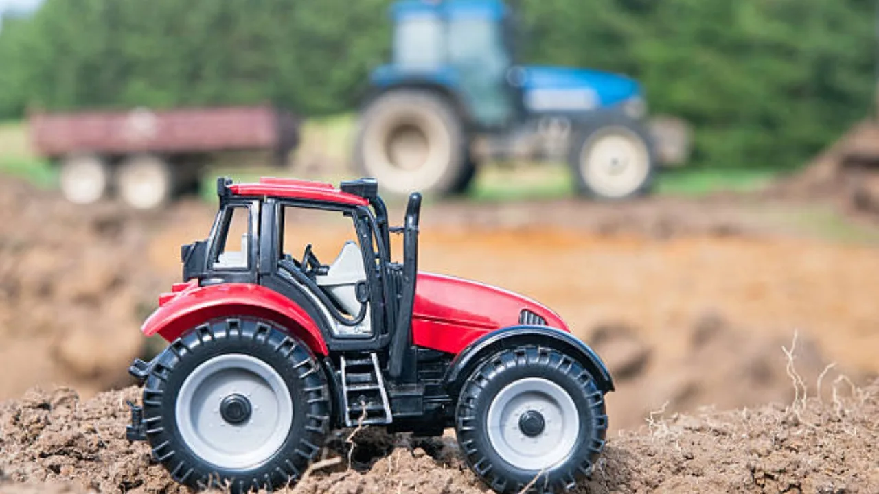 Tractor mic pe un teren agricol, cu un tractor mare în fundal.