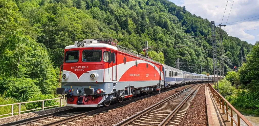 Tren modern circulând pe calea ferată în zona montană, cu vegetație abundentă în jur.