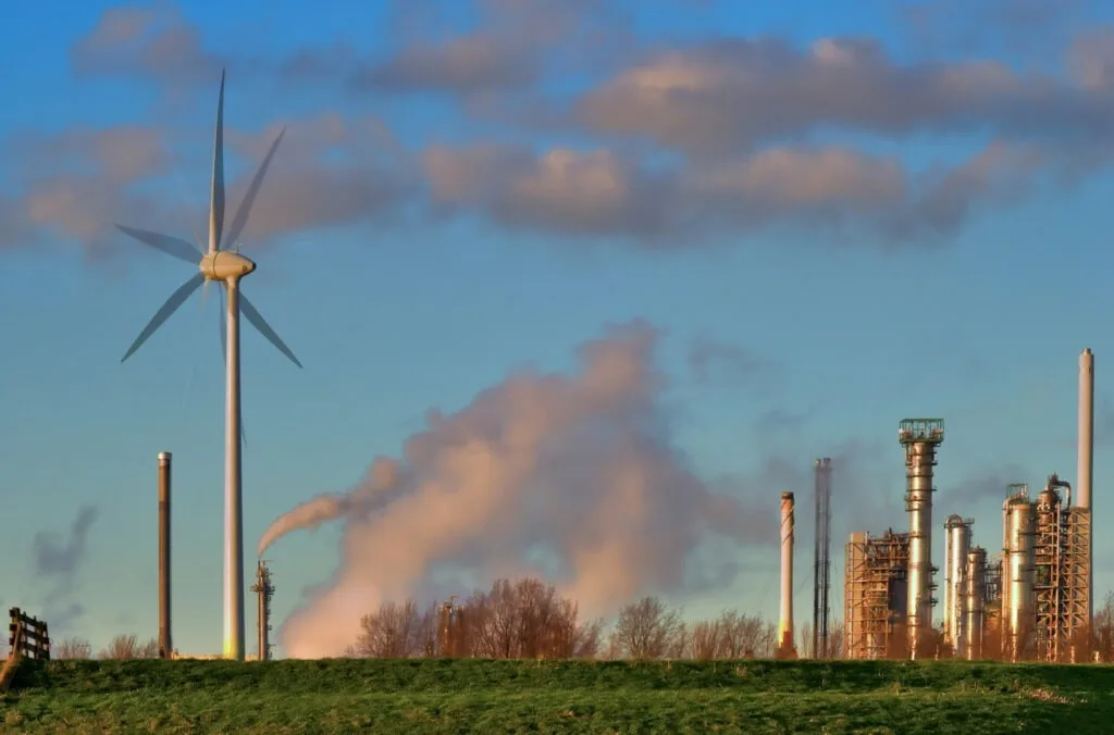 Peisaj industrial cu turbine eoliene și fum de la rafinării.