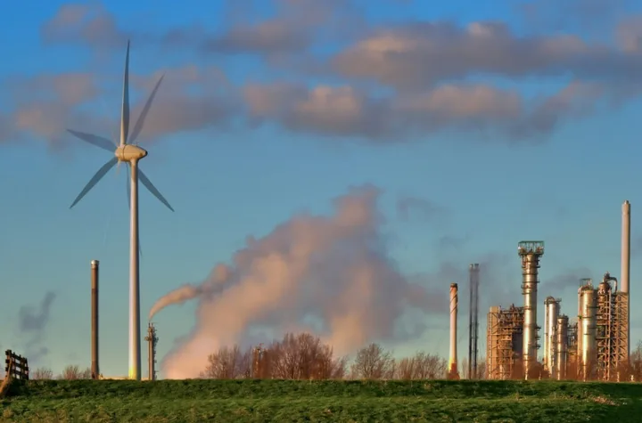 Peisaj industrial cu turbine eoliene și fum de la rafinării.
