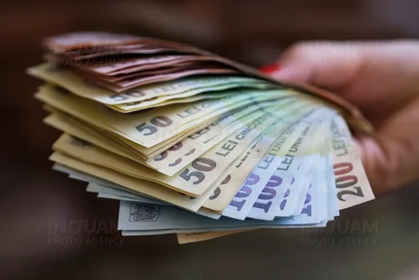 O mână ține bancnote românești de diferite valori, simbolizând presiunea financiară.