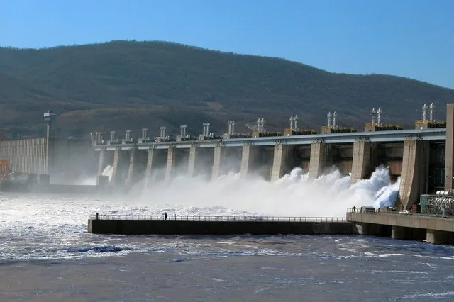 Baraj hidroelectric cu apă curgândă și peisaj montan în fundal.