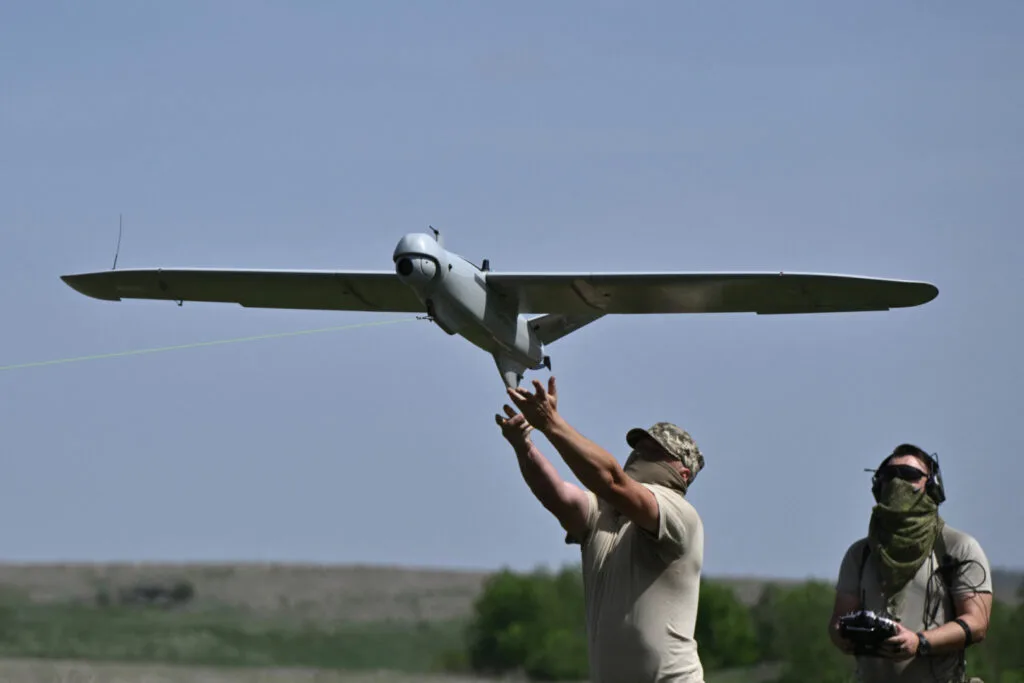 Doi bărbați lansează o dronă în aer, într-un peisaj deschis.