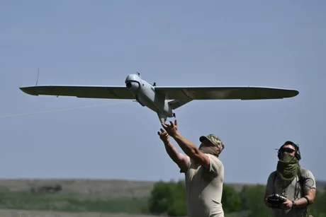 Doi bărbați lansează o dronă în aer, într-un peisaj deschis.
