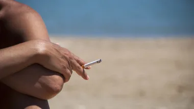 Un bărbat stând pe plajă, fumând o țigară, cu marea în fundal.