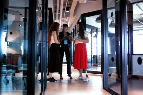 Trei femei discută într-un birou modern, înconjurate de sticlă și lumină naturală.