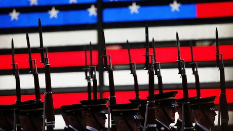 Soldați cu arme în fața unui fundal cu steagul american.