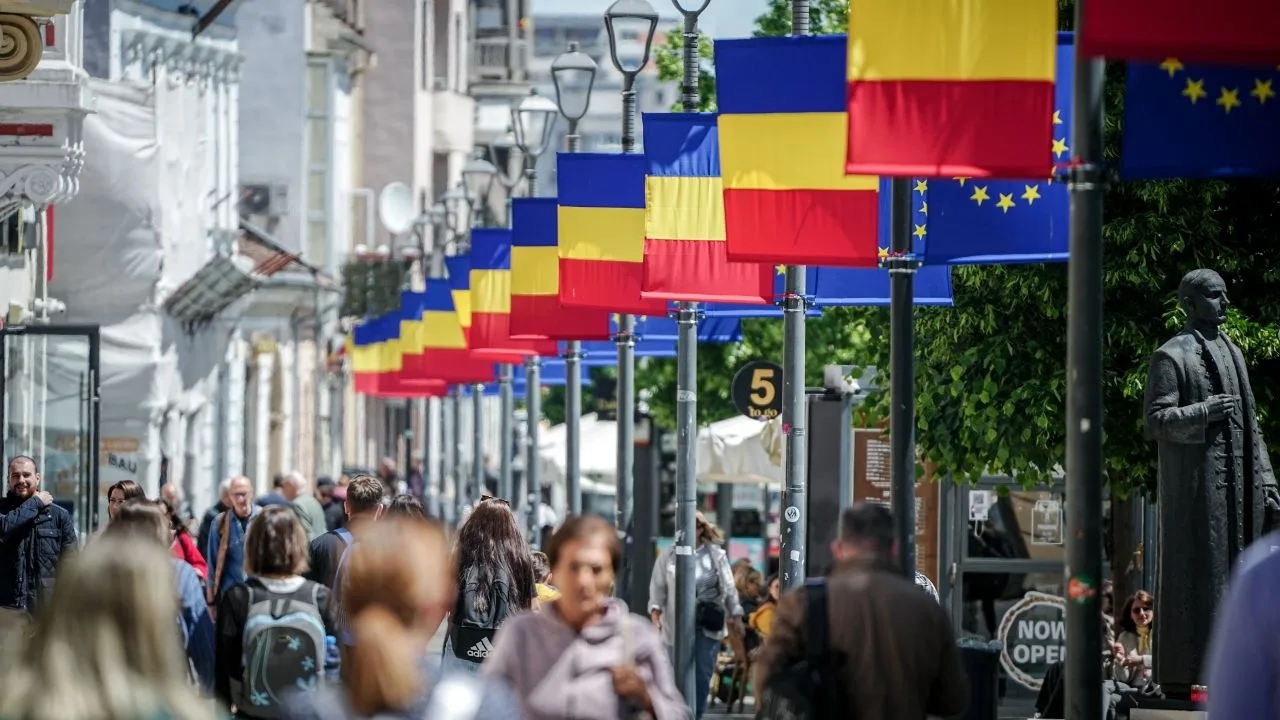 Stradă aglomerată cu oameni și steaguri românești fluturând la soare.