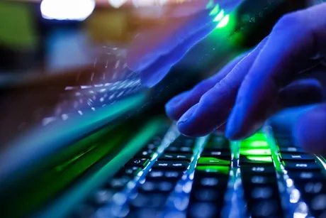 Mâini care tastează pe un keyboard iluminat, simbolizând spionajul cibernetic.