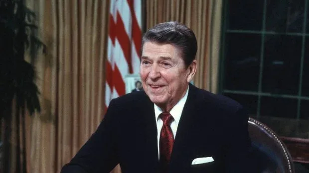 Ronald Reagan zâmbind în biroul Oval, cu drapelul în fundal.