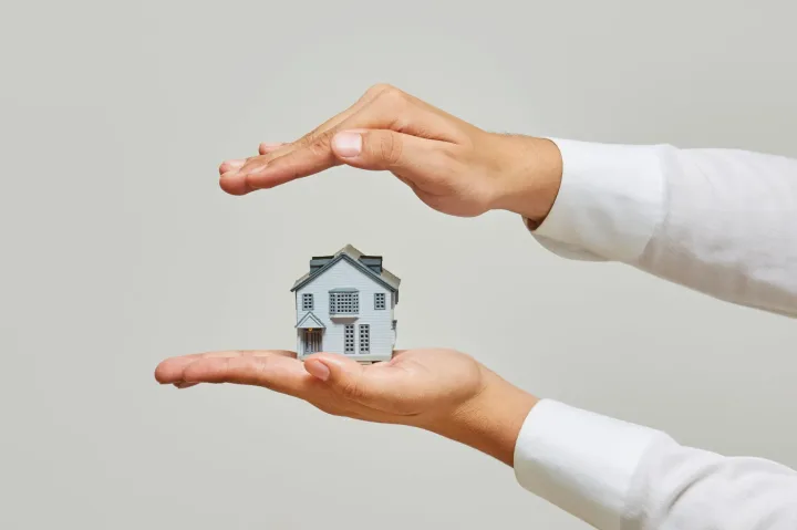 Două mâini protejează o miniatură a unei case, simbolizând asigurarea locuinței.