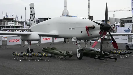 Dronă militară expusă, cu echipamente de armament alături.