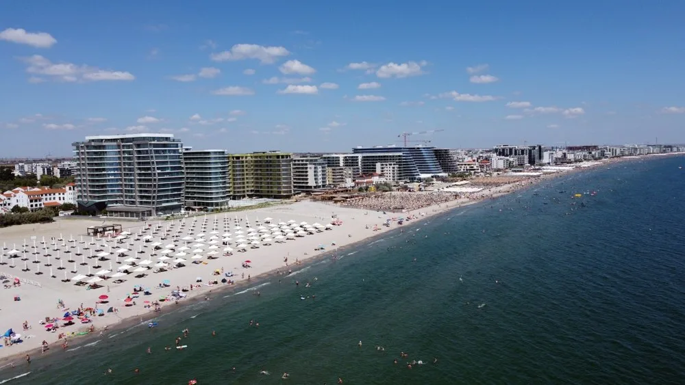 Plajă aglomerată pe litoral, cu hoteluri moderne în fundal și cer senin.